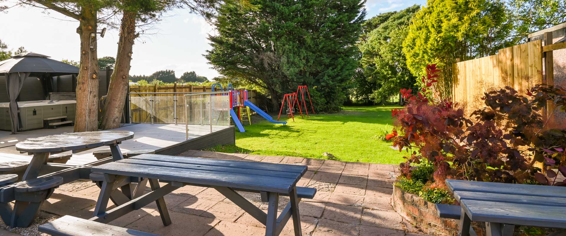 Paved outdoor patio with several picnic benches leading to a large lawn and hot tub.