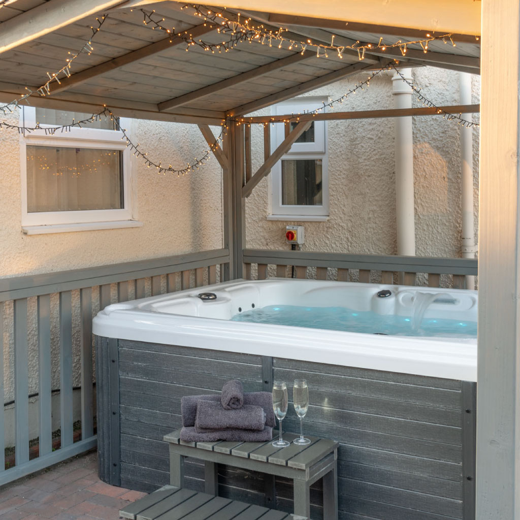 A luxury hot tub under a wooden gazebo with fairy lights and champagne flutes ready for guests.