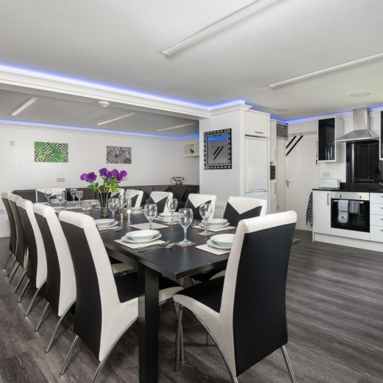 A long dining table with black and white chairs and blue LED ceiling lighting at Four Oaks House.