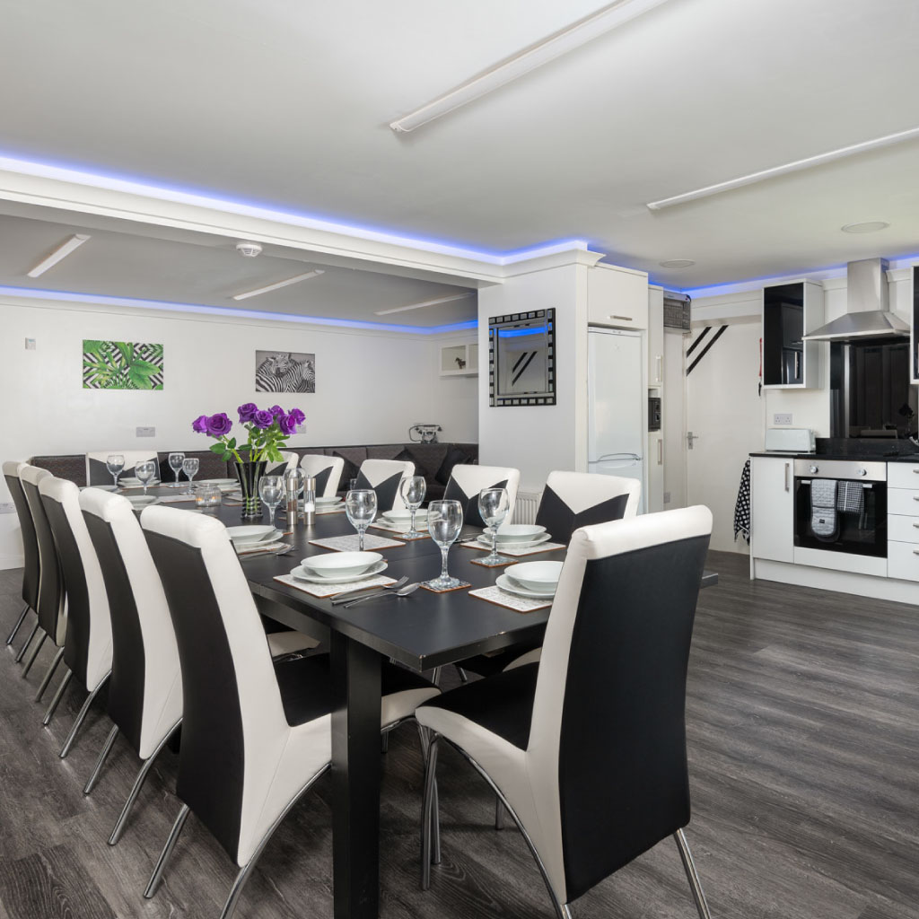 A long dining table with black and white chairs and blue LED ceiling lighting at Four Oaks House.